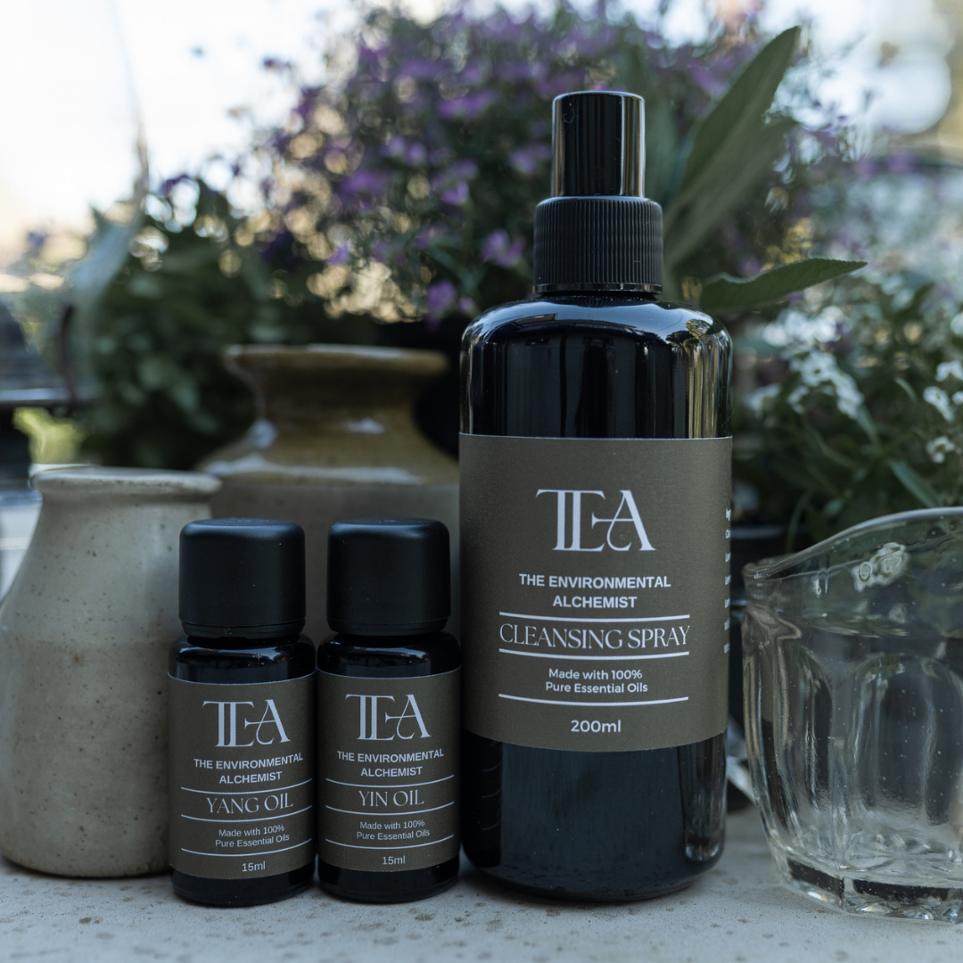 The image shows three bottles of products from "The Environmental Alchemist." There are two smaller bottles labeled "Yang Oil" and "Yin Oil," each 15ml, and a larger bottle labeled "Cleansing Spray," 200ml. The bottles are set against a background of plants and pottery.