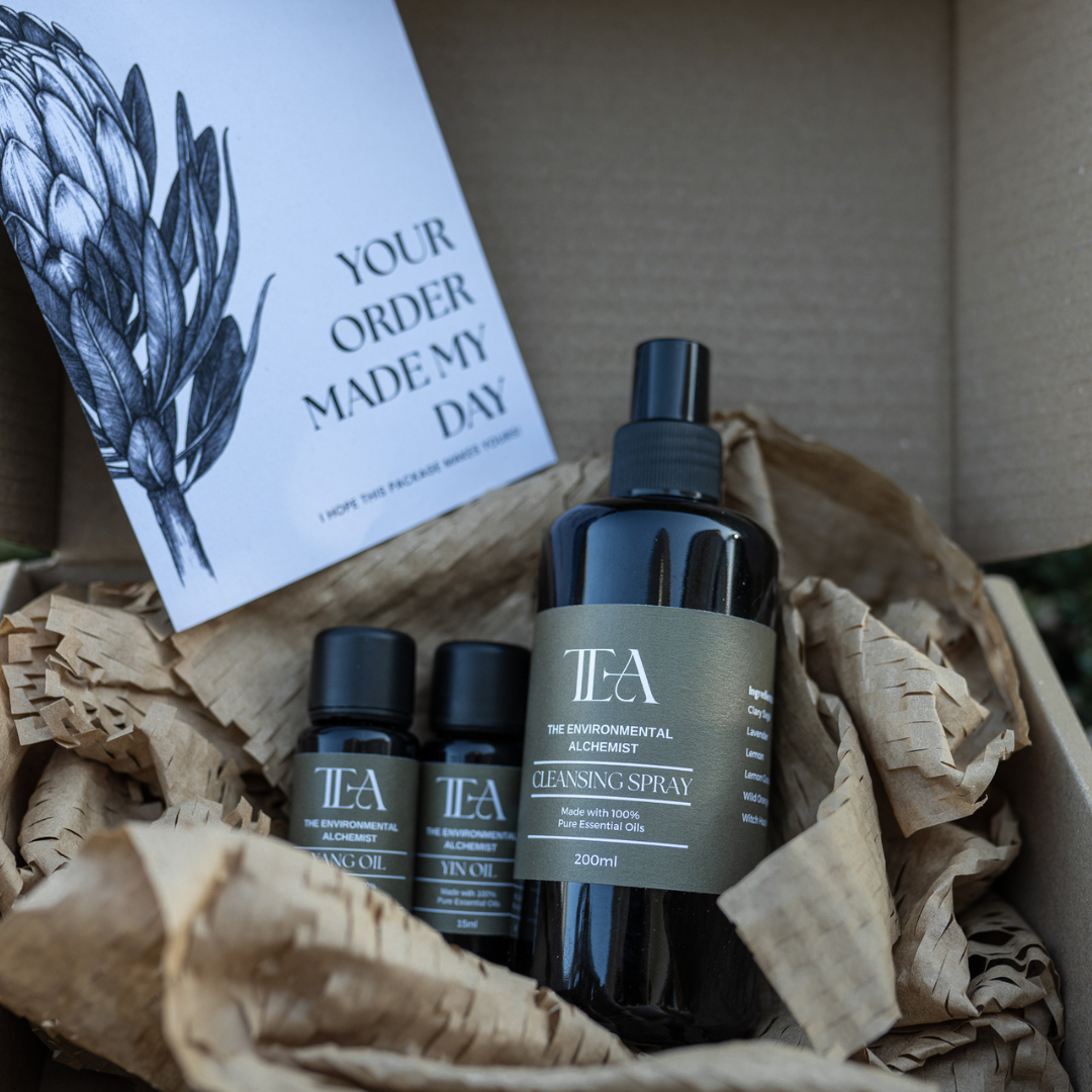 The image shows three bottles of products from "The Environmental Alchemist." There is a large bottle labeled "Cleansing Spray" and two smaller bottles labeled "Yang Oil" and "Yin Oil." The bottles are placed on a surface with plants and pottery in the background.