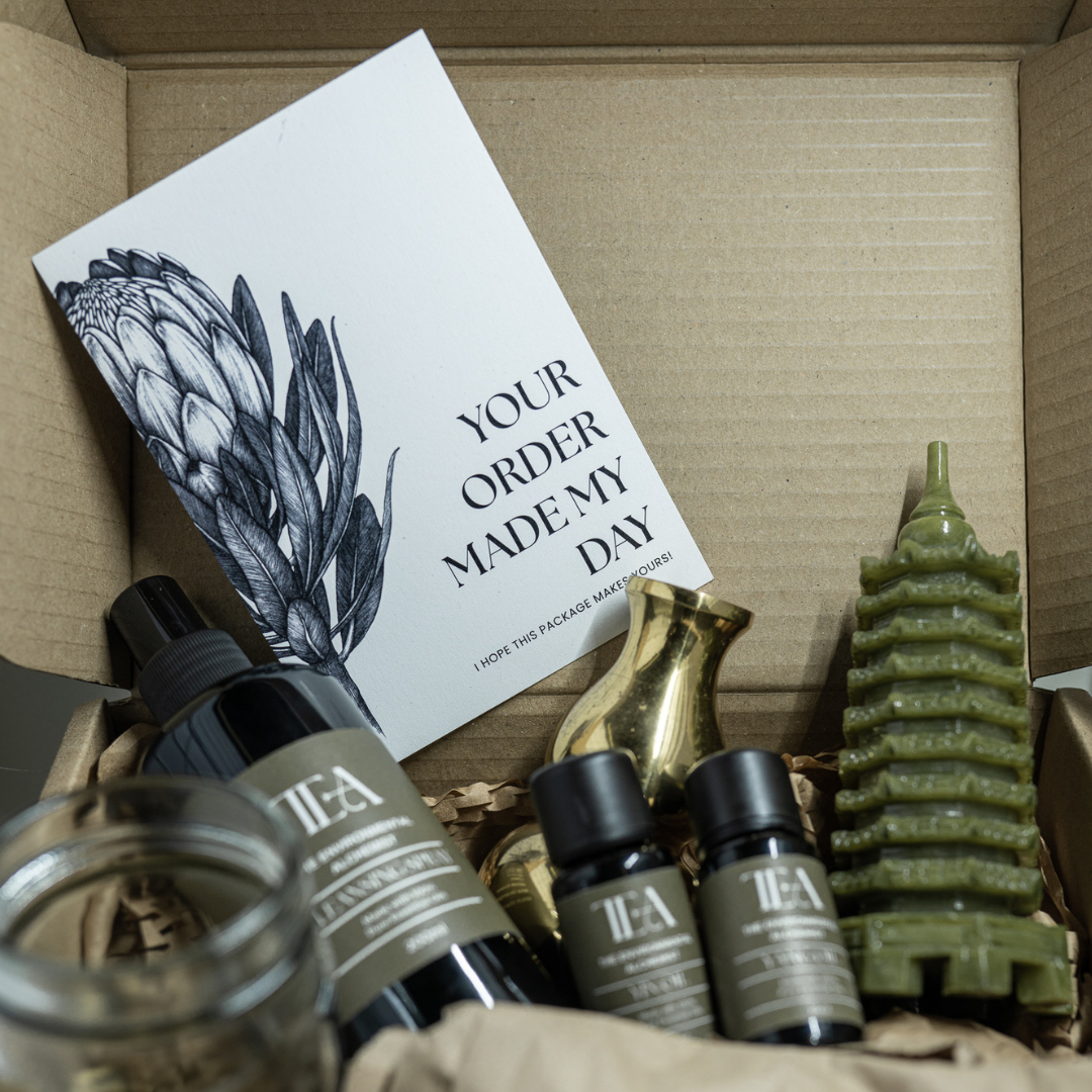 The image shows an open cardboard box containing various items. Inside, there is a card with the text "YOUR ORDER MADE MY DAY" and an illustration of an artichoke. The box also contains a green candle shaped like a pagoda, a glass jar, and several bottles labeled "TEA" with different products. Additionally, there are two small gold-colored vases.
