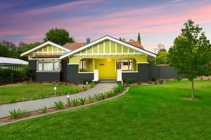 A single-story house with a vibrant yellow and dark gray exterior. The house features a gabled roof with red tiles and white trim. There is a well-maintained lawn and a curved pathway leading to the entrance. The sky is a colorful mix of pink and purple hues, suggesting sunset or sunrise.