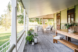 A spacious porch with wooden flooring and a white ceiling. There are several potted plants along the railing. A long wooden table with benches is on the right side, and there are chairs in the background. The porch overlooks a grassy area with trees.
