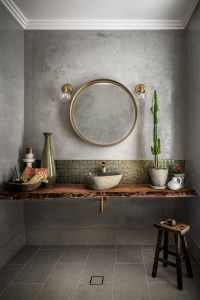 A stylish bathroom features a round mirror with a gold frame, mounted on a textured gray wall. Below the mirror is a wooden countertop with a stone sink and gold faucet. The backsplash is made of small green tiles. On the counter, there are decorative items including a tall green vase, a small potted cactus, a bowl with towels, and a small white pitcher. Two wall-mounted light fixtures with clear glass bulbs flank the mirror.